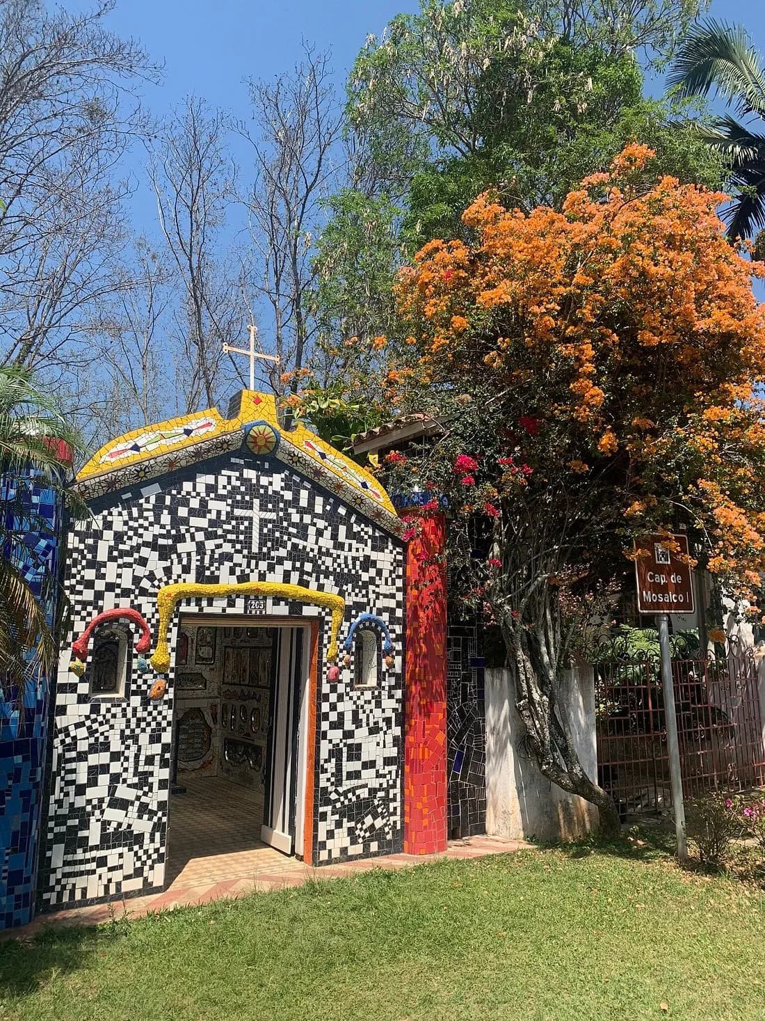 Interior da capelinha coberto de mosaico: paredes com imagens de santos, Cristo no frontão e altar de cacos coloridos
