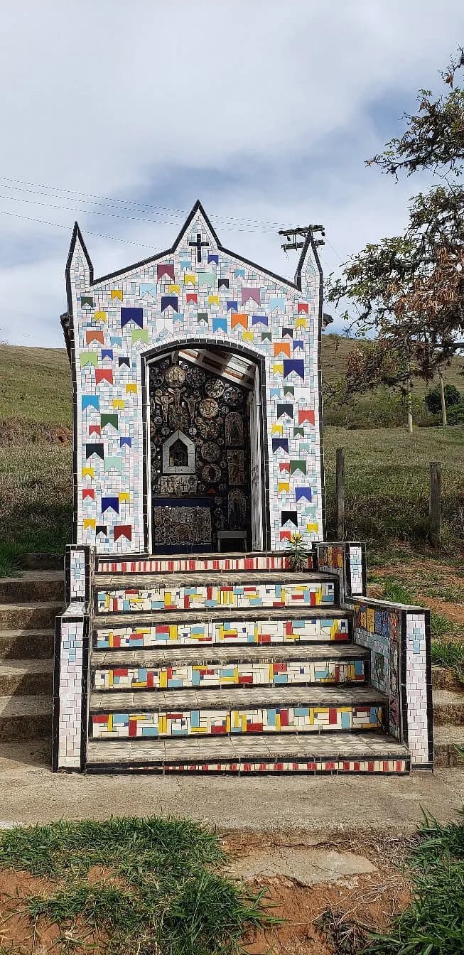 Capelinha de mosaico do Paiol, com fachada de bandeirinhas coloridas e escadaria de caco no meio do pasto