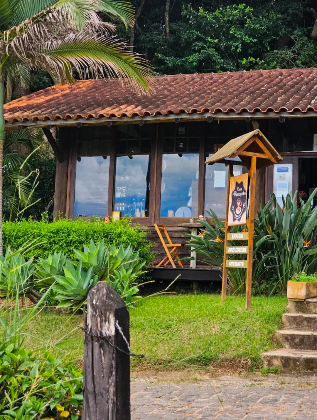 Casa de madeira do Dona Mariquinha com telhado de barro, janelões de vidro e a placa de empório, cafés especiais e artesanato no gramado da frente