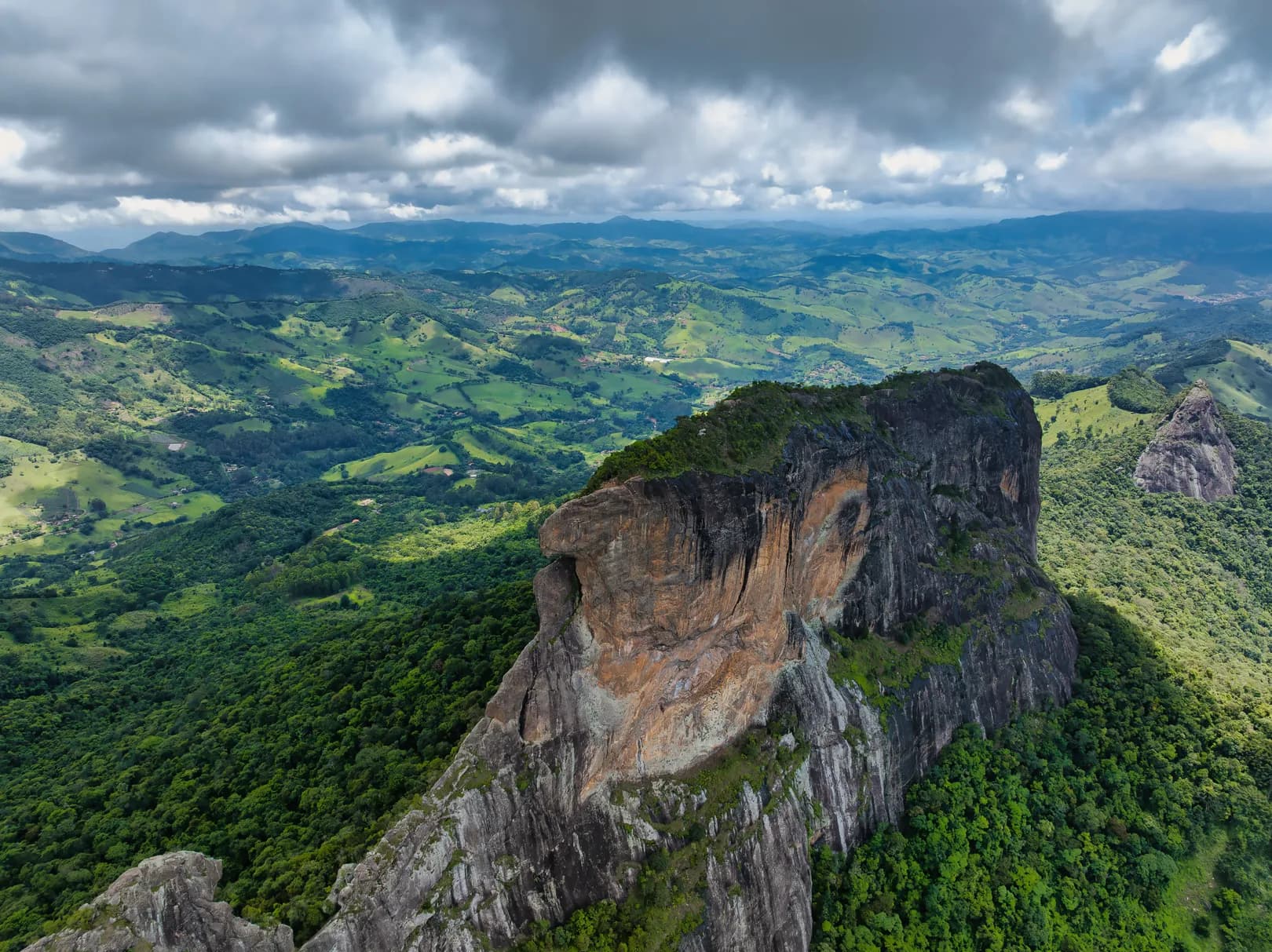 Vista aérea do paredão da Pedra do Baú sobre a mata, com o vale verde e as montanhas ao fundo