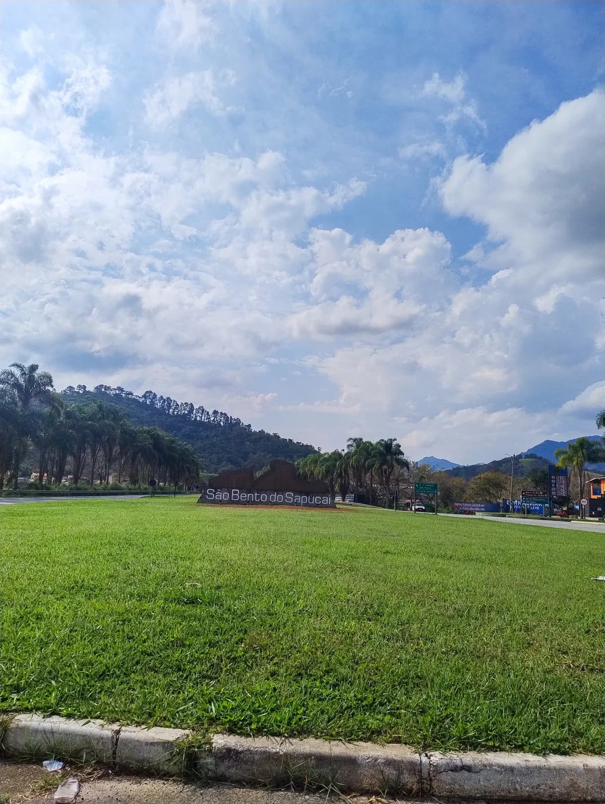 Monumento com o nome São Bento do Sapucaí e a silhueta das montanhas, sobre um gramado na entrada da cidade, com morros ao fundo
