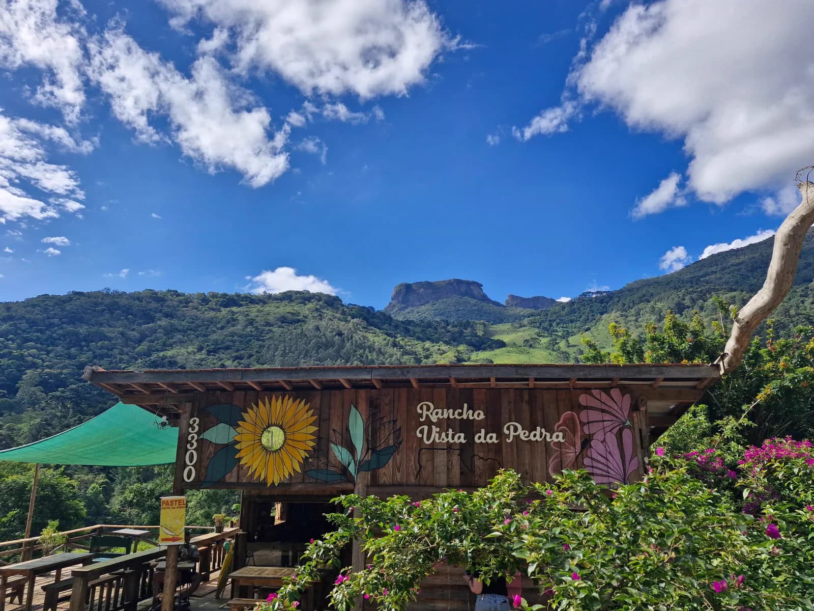 Quiosque de madeira com o número 3300, um girassol pintado e o nome Rancho Vista da Pedra na parede, com a Pedra do Baú ao fundo