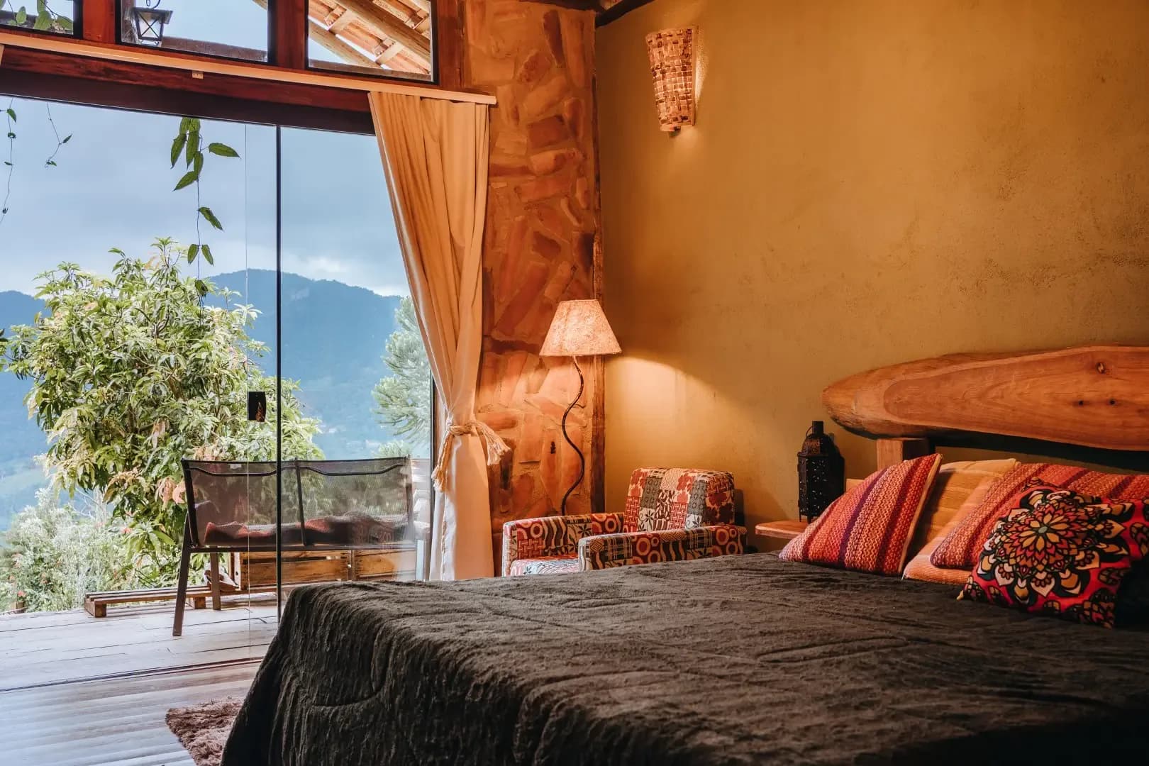Quarto da Cabana Ametista com cama de casal, cabeceira de tronco maciço, parede de barro e porta de vidro para a varanda com vista da montanha