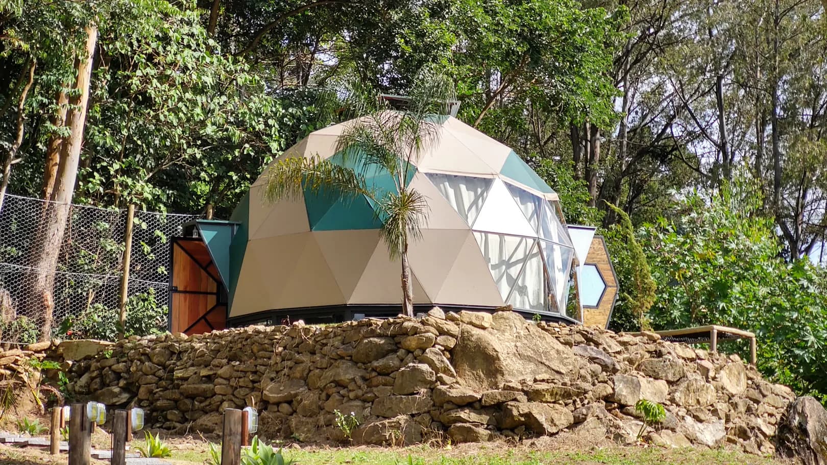 Domo geodésico Colmeia com painéis bege e verdes e fachada de vidro, sobre muro de pedra em meio à mata