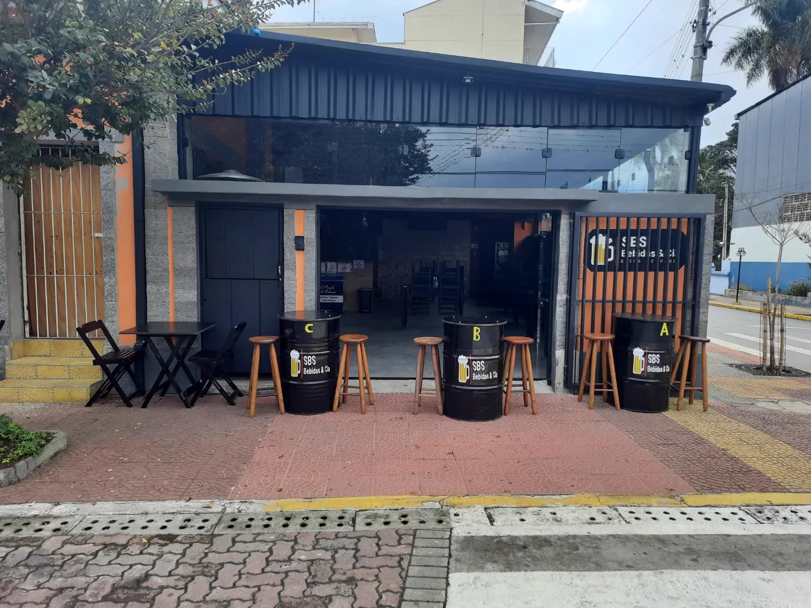 Fachada preta do SBS Bebidas & Cia de dia, com vitrine de vidro, portão laranja e três tonéis marcados A, B e C com banquetas de madeira na calçada