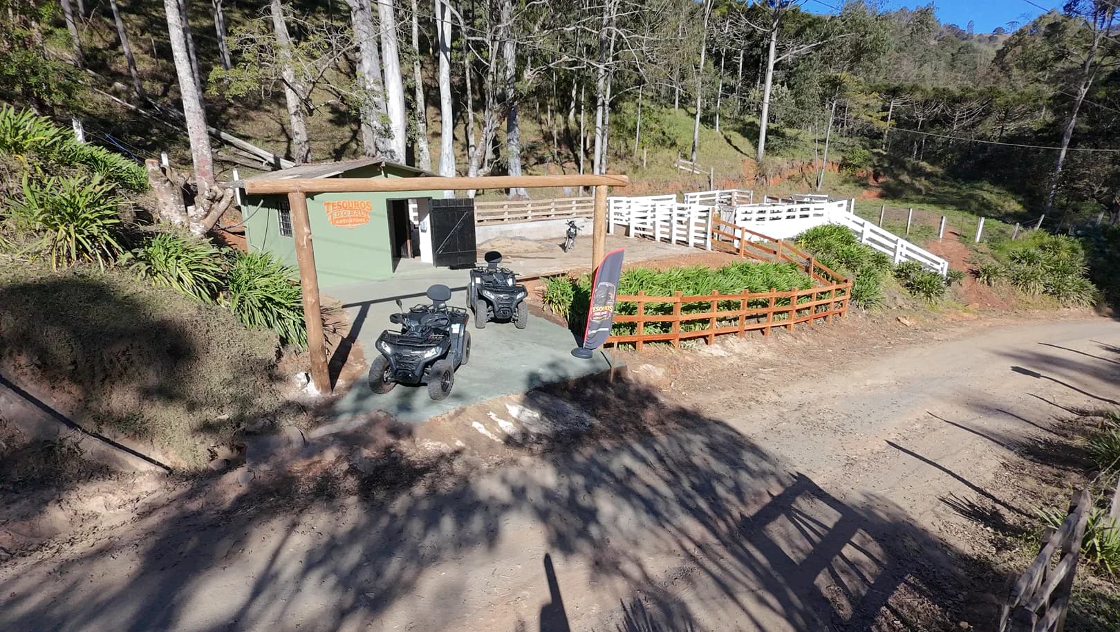 Dois quadriciclos estacionados sob um pórtico de madeira, em frente a uma casa verde com placa de Tesouros do Baú Adventure, na beira de uma estrada de terra