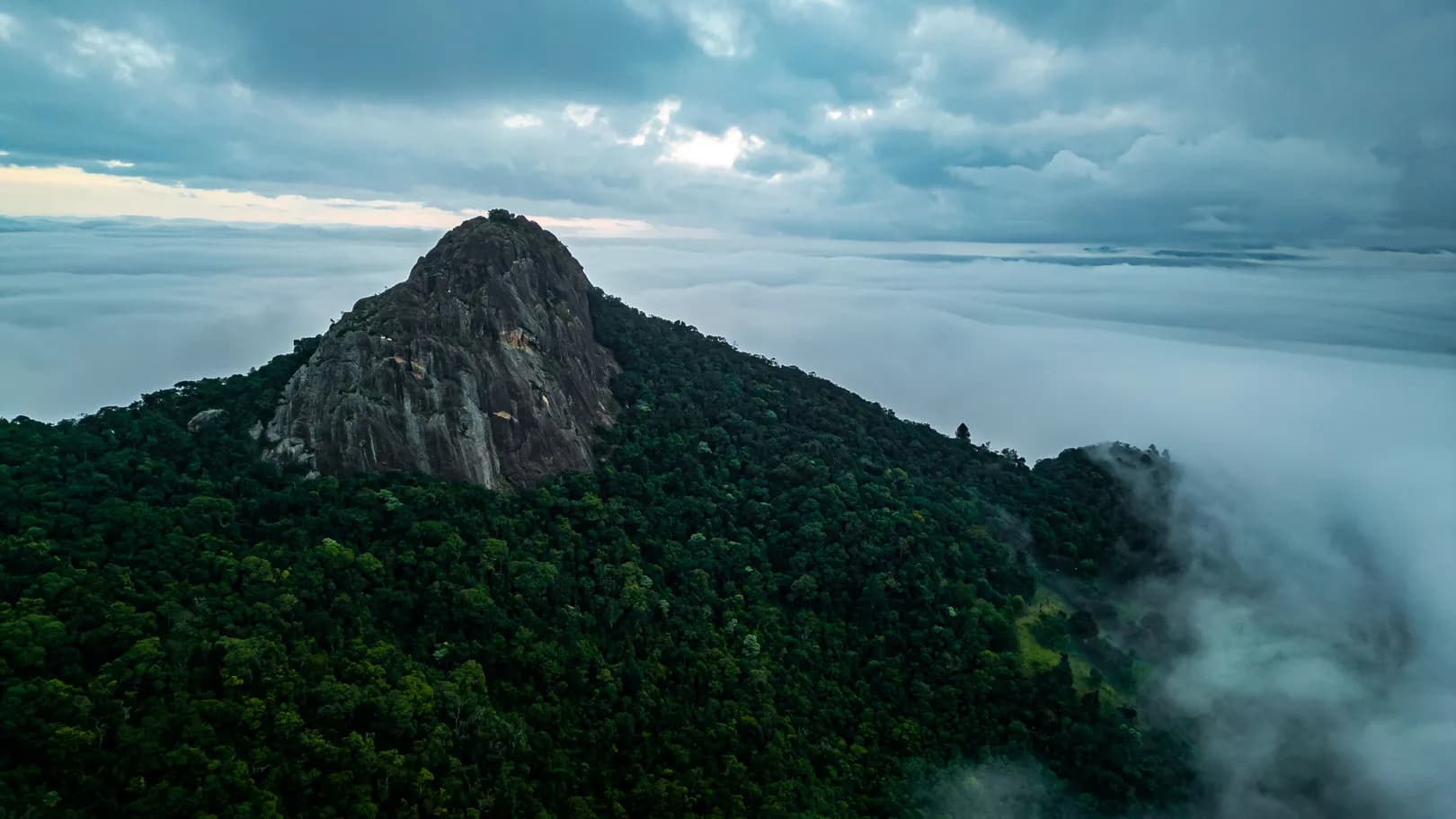 Monólito do complexo do Baú visto de cima, sob céu carregado, cercado de mata e acima de um mar de nuvens