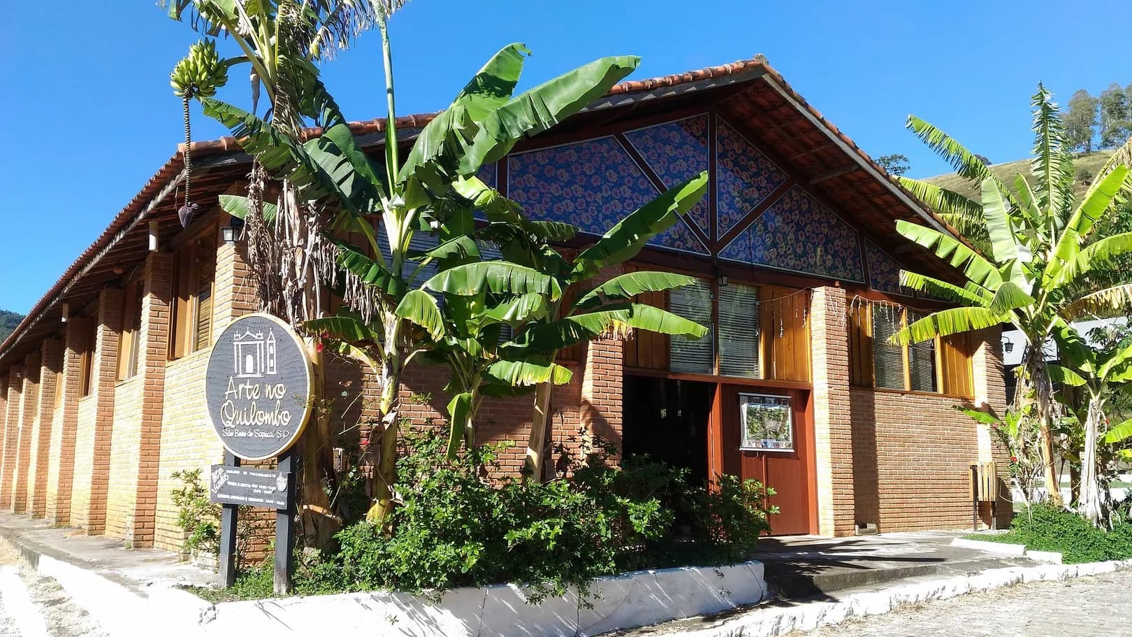 Fachada de tijolo e madeira do Arte no Quilombo, em São Bento do Sapucaí, com telhado de telha, bananeiras e a placa redonda na entrada