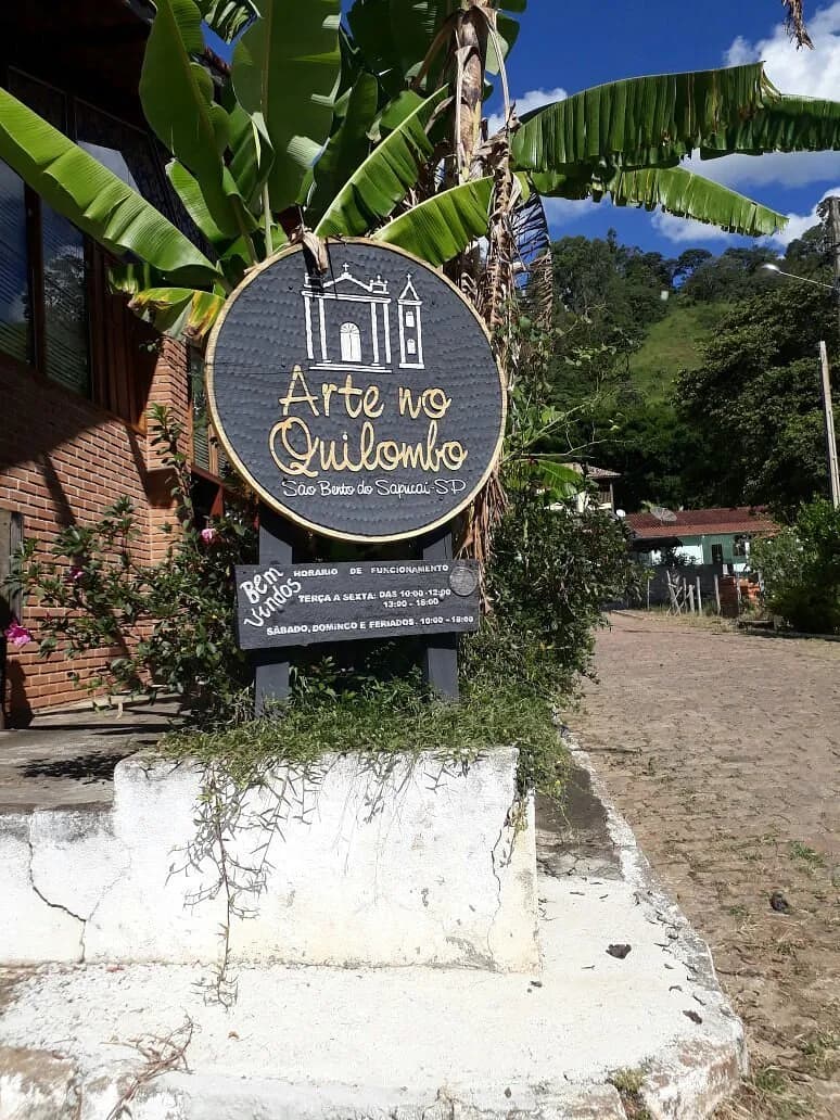 Placa redonda de madeira do Arte no Quilombo, com a silhueta de uma capela e os horários de funcionamento