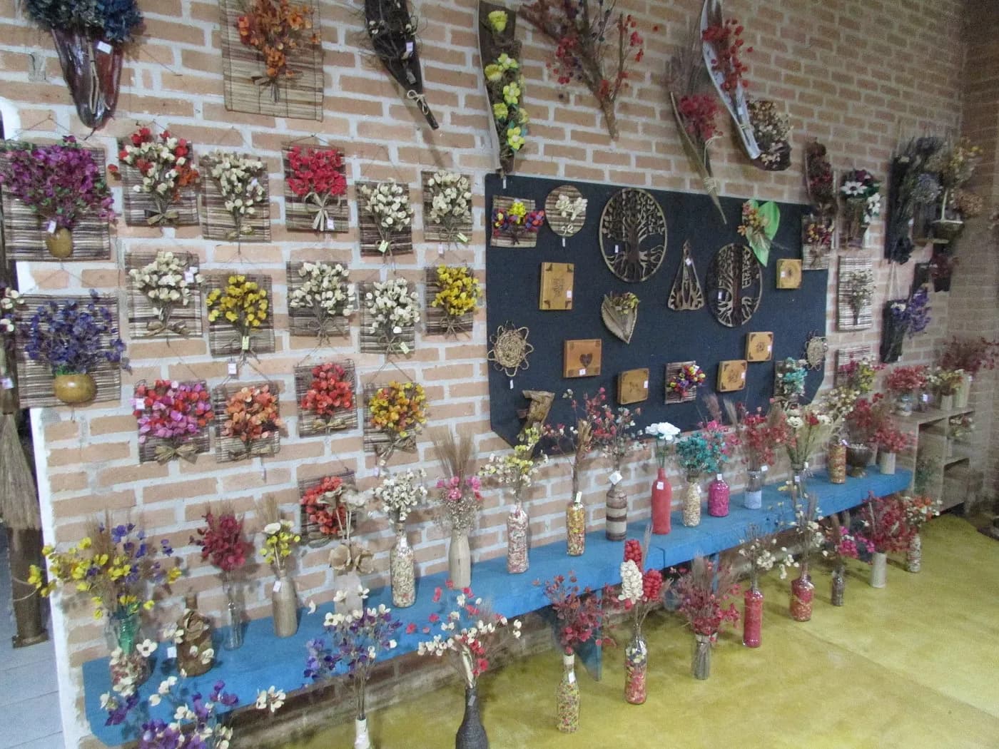 Parede de tijolo coberta de arranjos de flores secas, com garrafas e vasos enfileirados na mesa embaixo