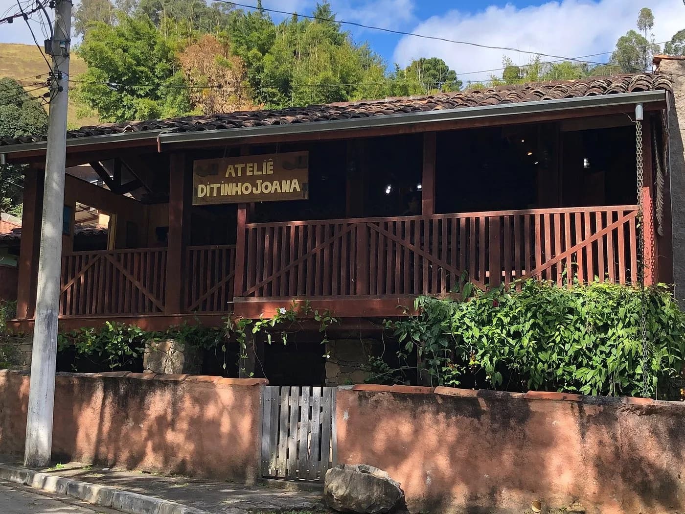 Fachada do Ateliê Ditinho Joana em São Bento do Sapucaí: casa de madeira avermelhada com varanda, placa entalhada e muro rosado