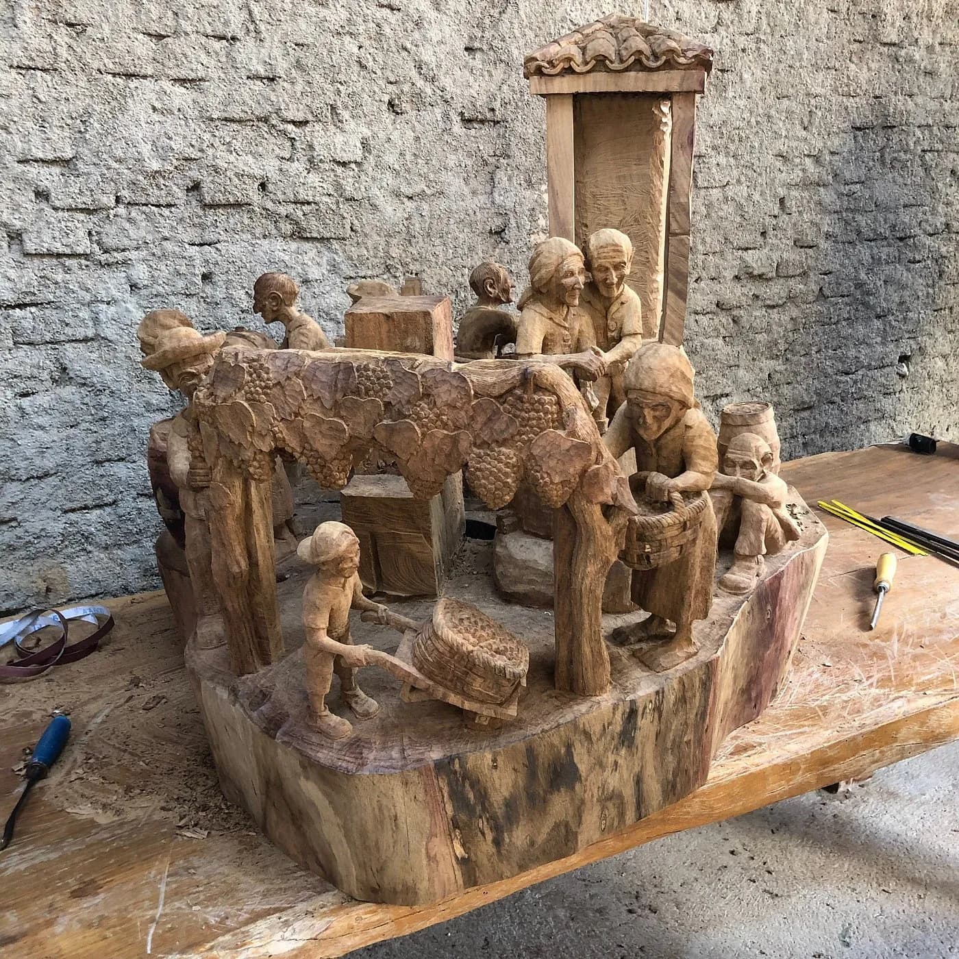Escultura em produção sobre a bancada do ateliê, com cena de colheita sob a parreira e as goivas ao lado