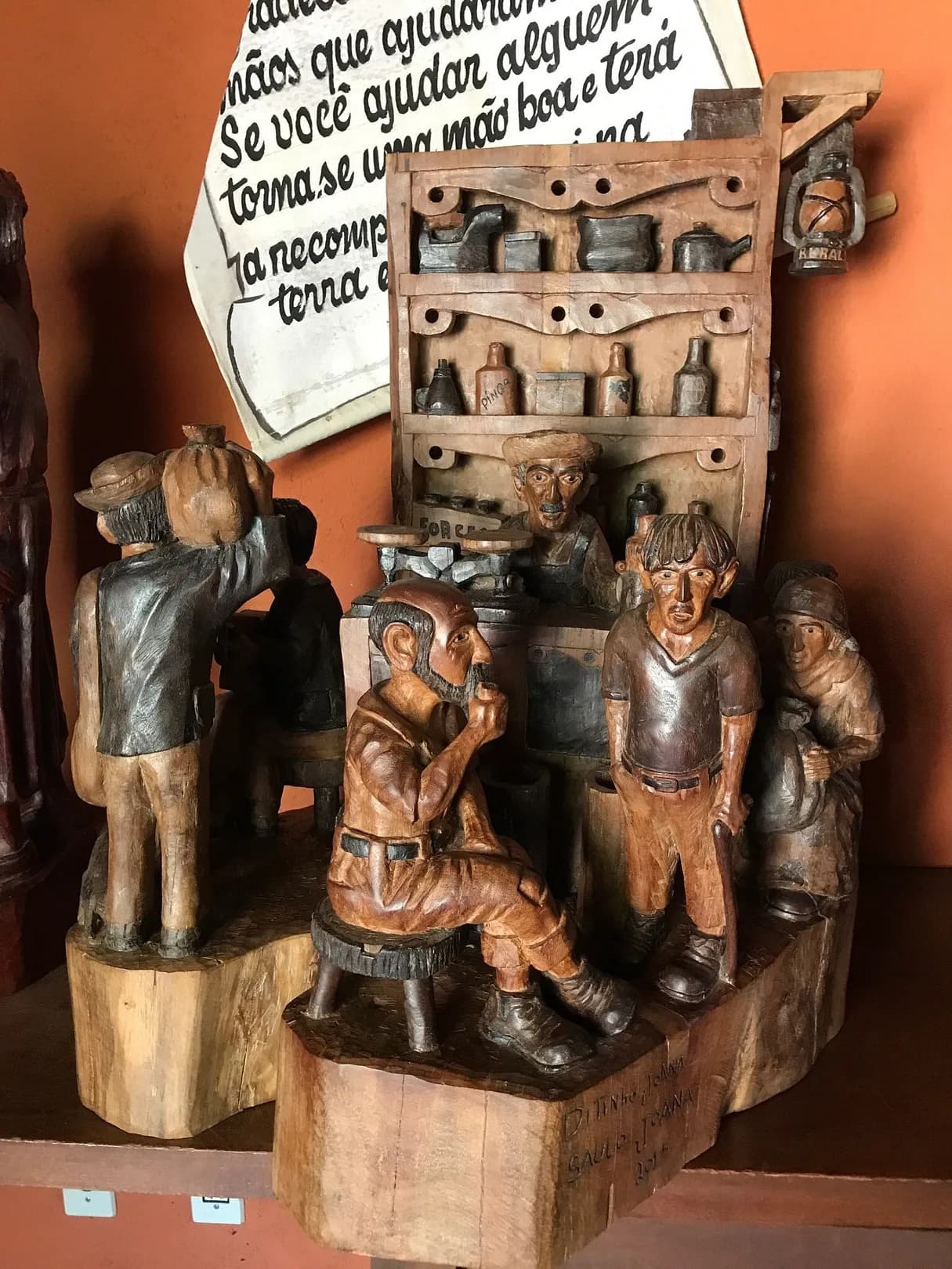 Conjunto de esculturas de madeira representando uma venda antiga, com figuras sentadas e prateleiras de garrafas