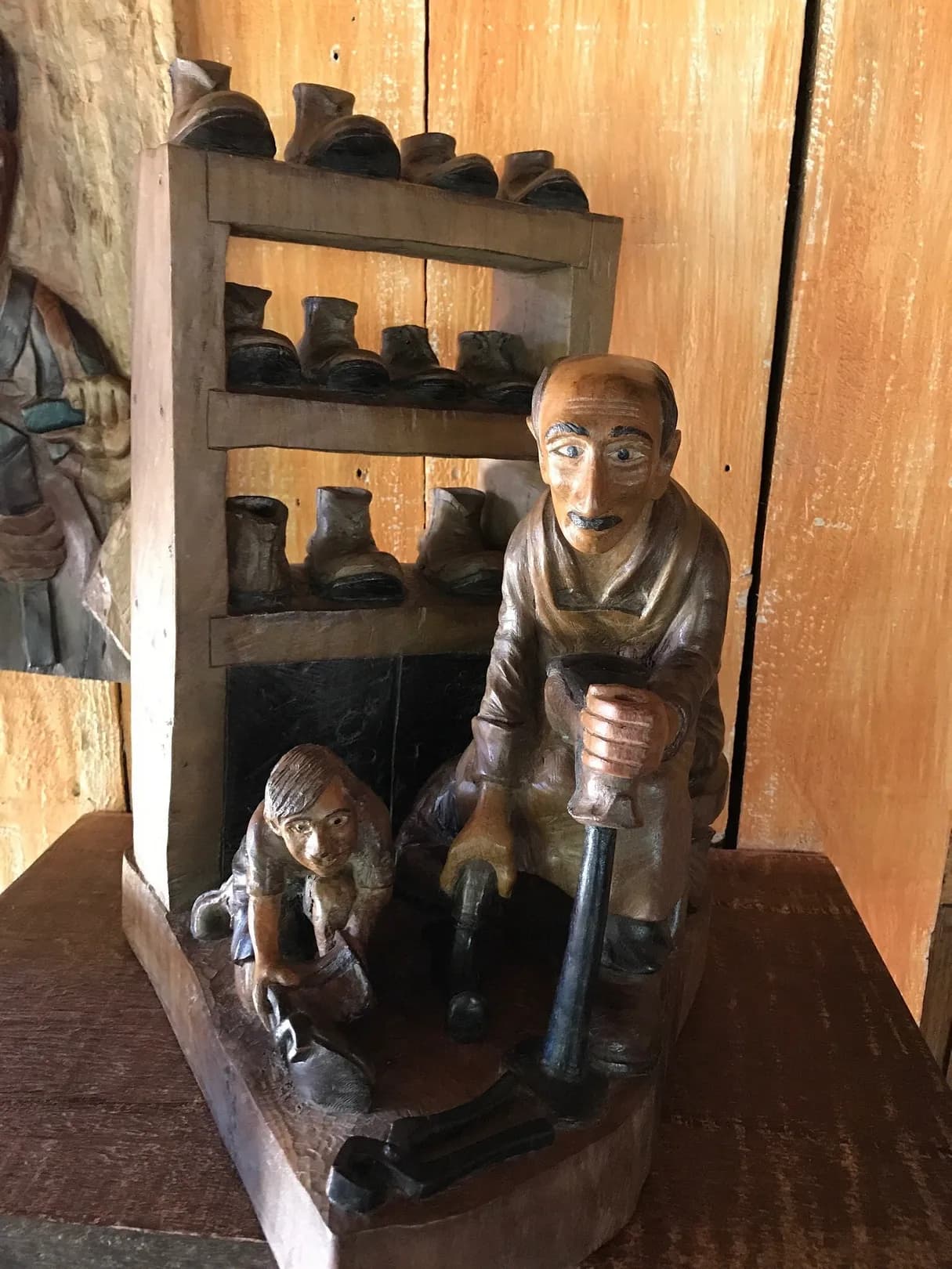 Escultura de um sapateiro com uma criança ao lado, diante da prateleira de botinas talhadas em madeira