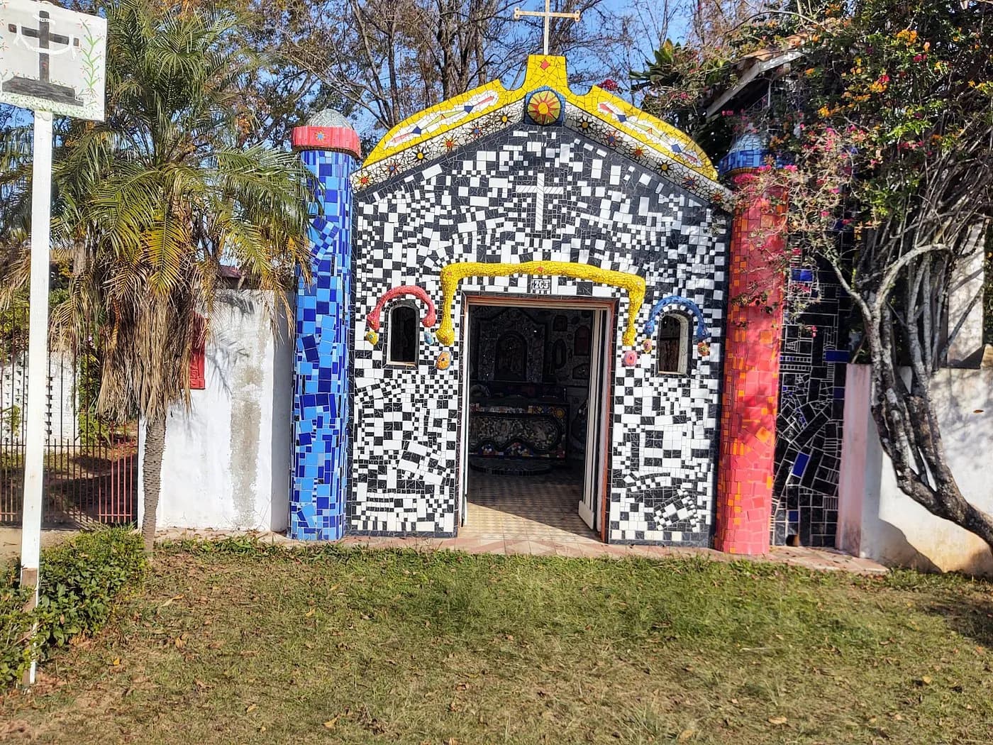 Capelinha de mosaico em São Bento do Sapucaí, revestida de caco preto e branco, com colunas azul e vermelha, frontão amarelo e porta aberta
