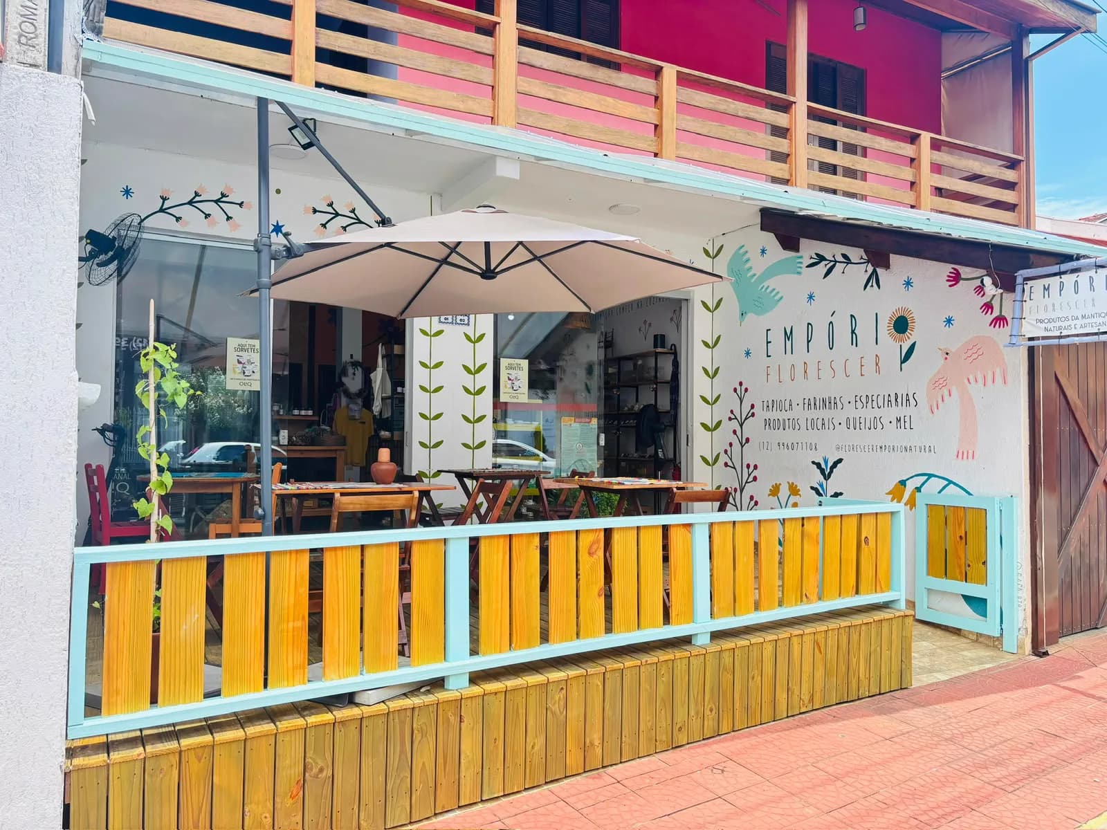 Fachada do Empório Florescer em São Bento do Sapucaí, com deck de madeira, mesas sob guarda-sol e mural de flores e pássaros