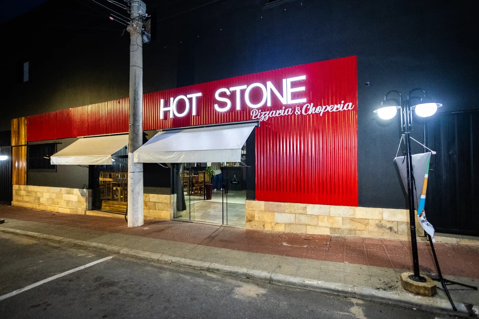 Fachada da Hot Stone Pizzaria & Choperia à noite, com o letreiro branco iluminado sobre painel vermelho canelado e os toldos claros da entrada