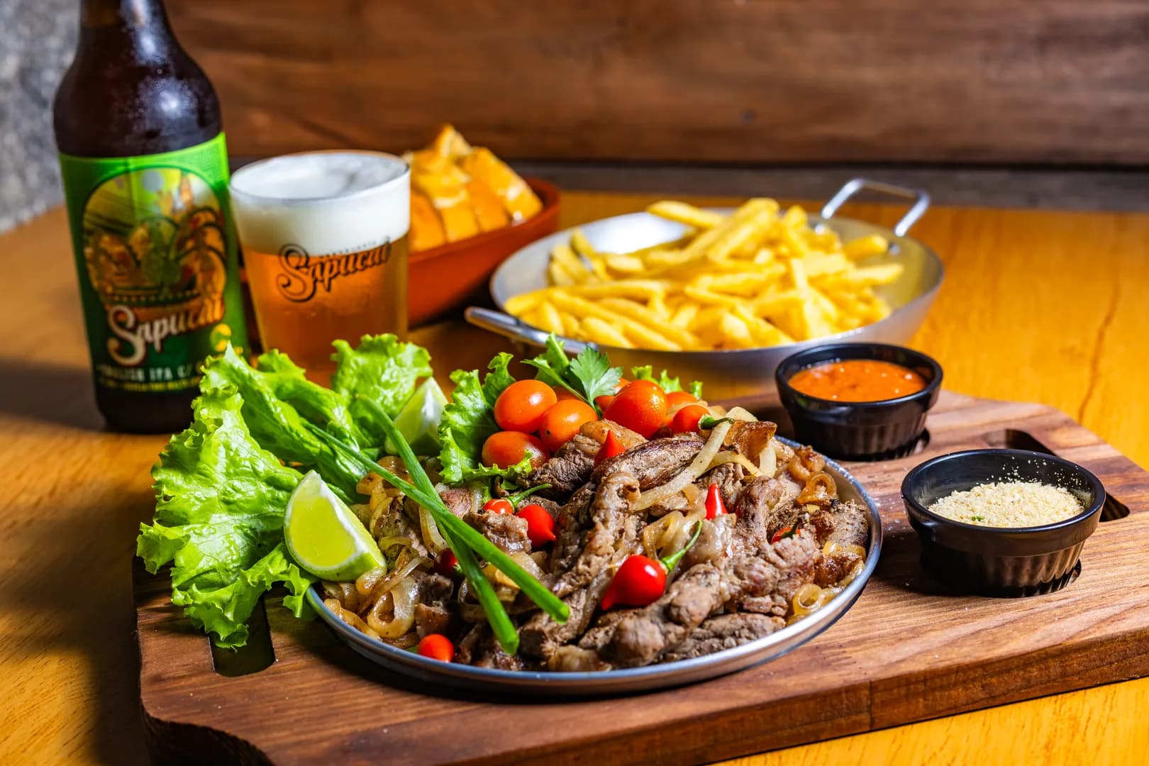 Tábua com carne fatiada, cebola e tomatinhos, fritas na frigideira, farofa, molho e um chope da Cervejaria Sapucaí ao lado