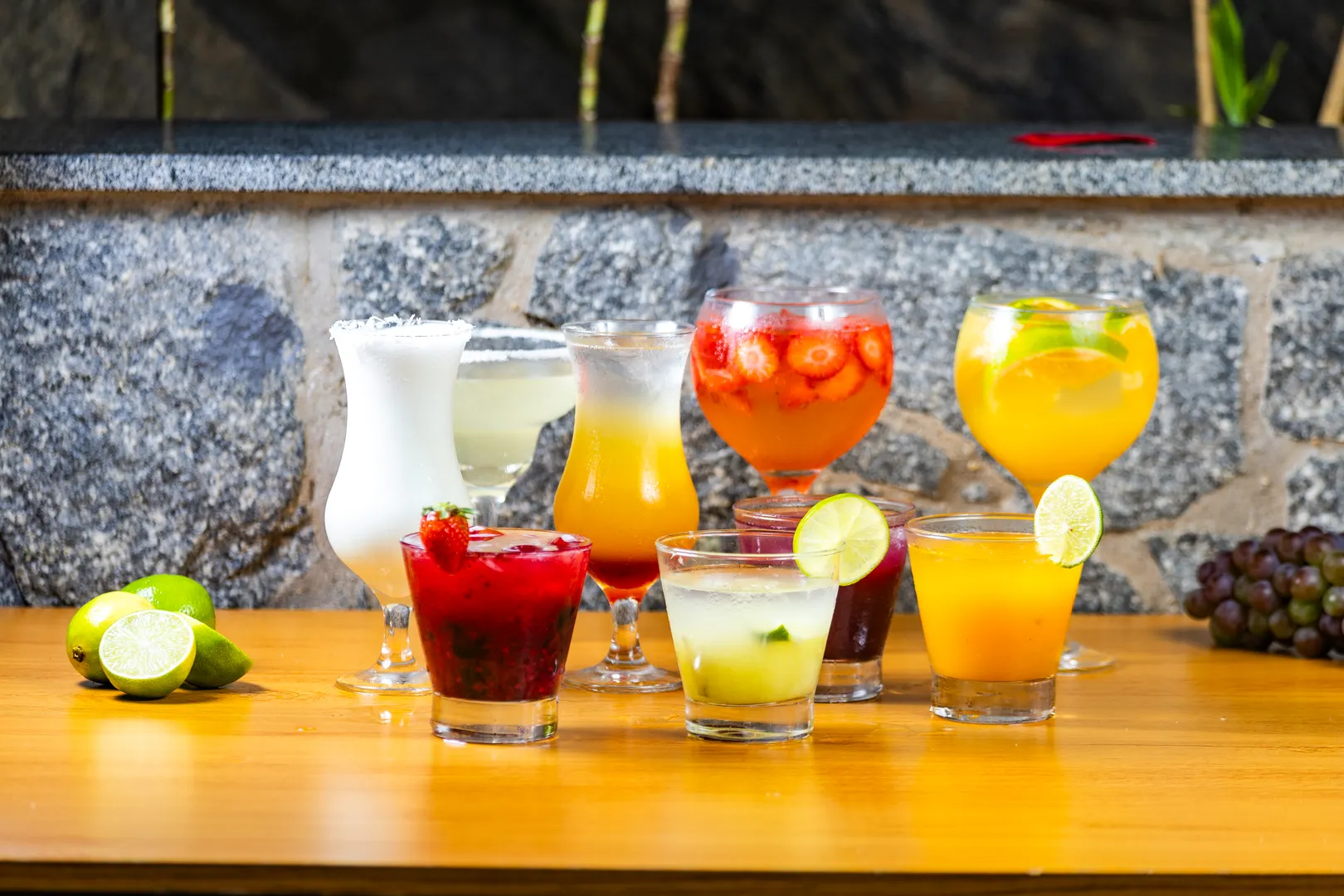 Fileira de drinques coloridos no balcão de madeira da Hot Stone, com limões e uvas ao lado e a parede de pedra atrás