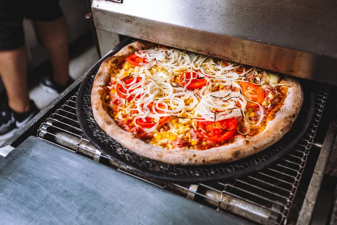 Pizza de tomate, cebola e milho saindo na chapa preta pela esteira do forno da Hot Stone