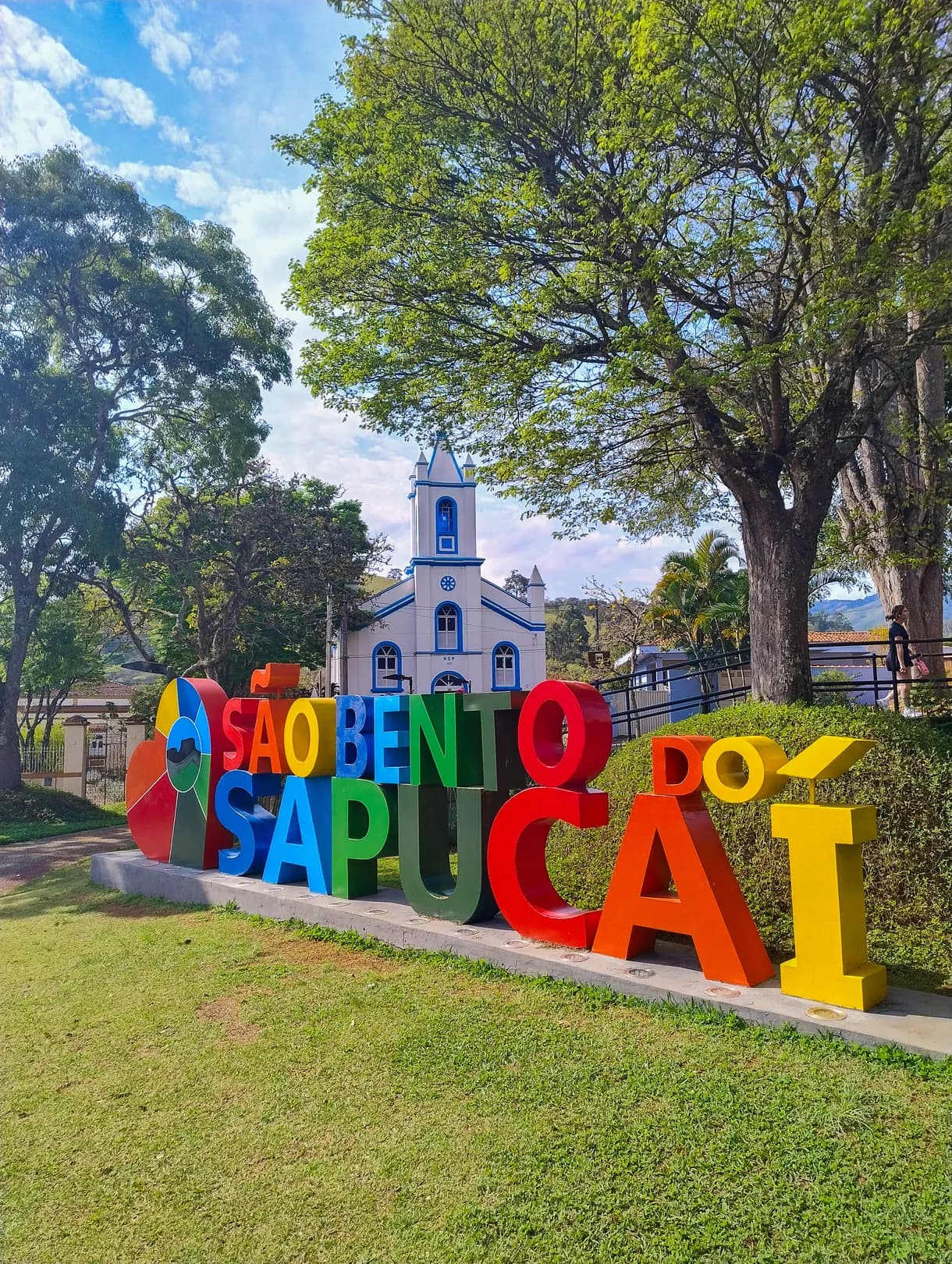 Letreiro colorido de São Bento do Sapucaí no gramado da praça, com a Igreja Nossa Senhora do Rosário, branca e azul, logo atrás