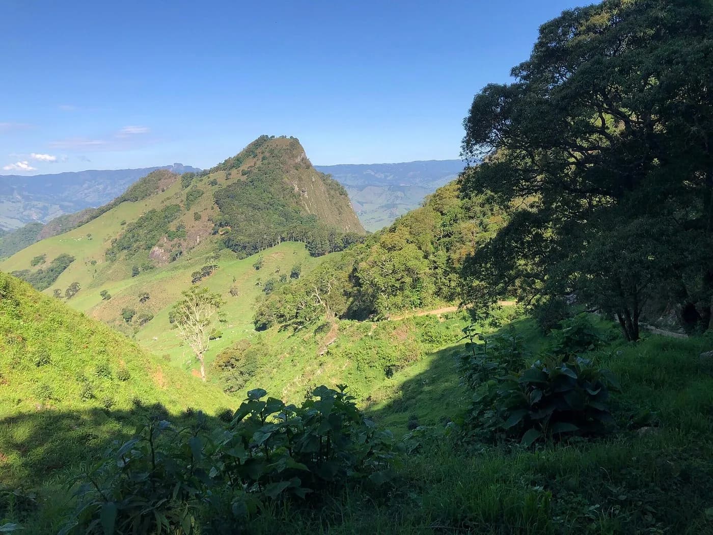 Pedra da Balança, morro pontiagudo de mata e pasto sobre o vale, com estrada de terra na encosta e a Serra da Mantiqueira ao fundo, em São Bento do Sapucaí