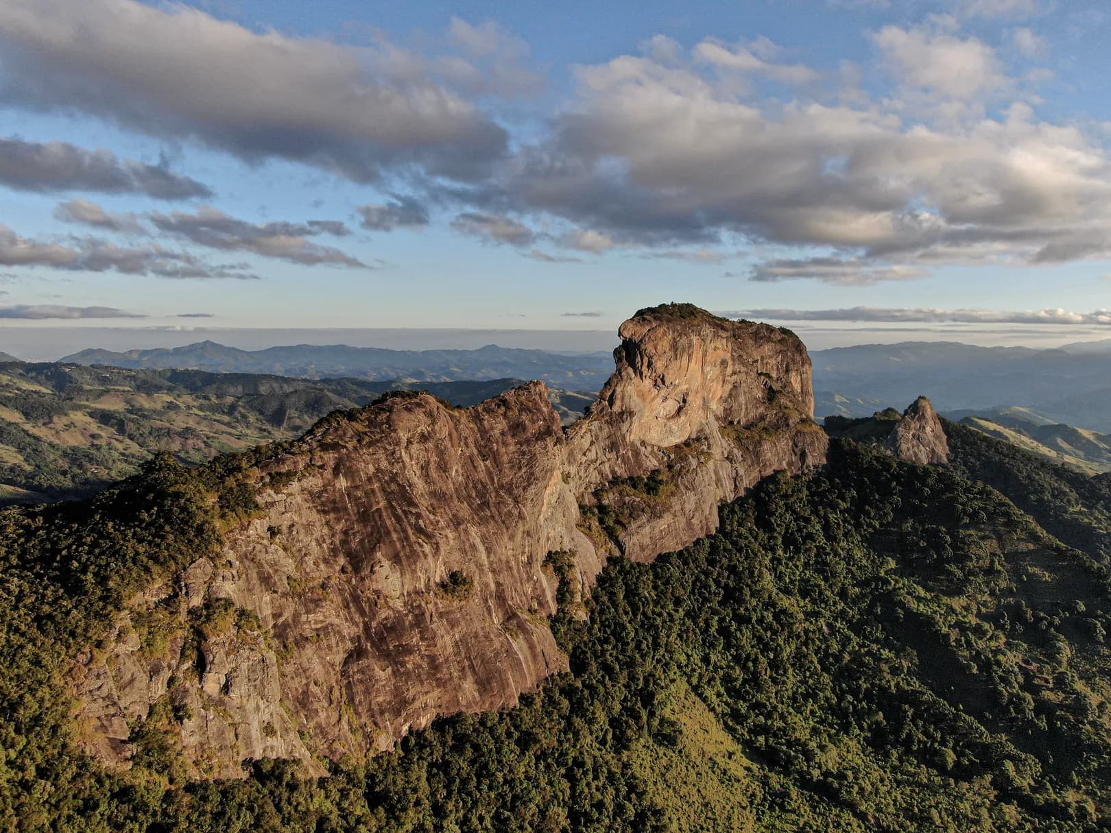 Vista aérea do paredão da Pedra do Baú à luz do fim de tarde, com a Serra da Mantiqueira ao fundo, em São Bento do Sapucaí