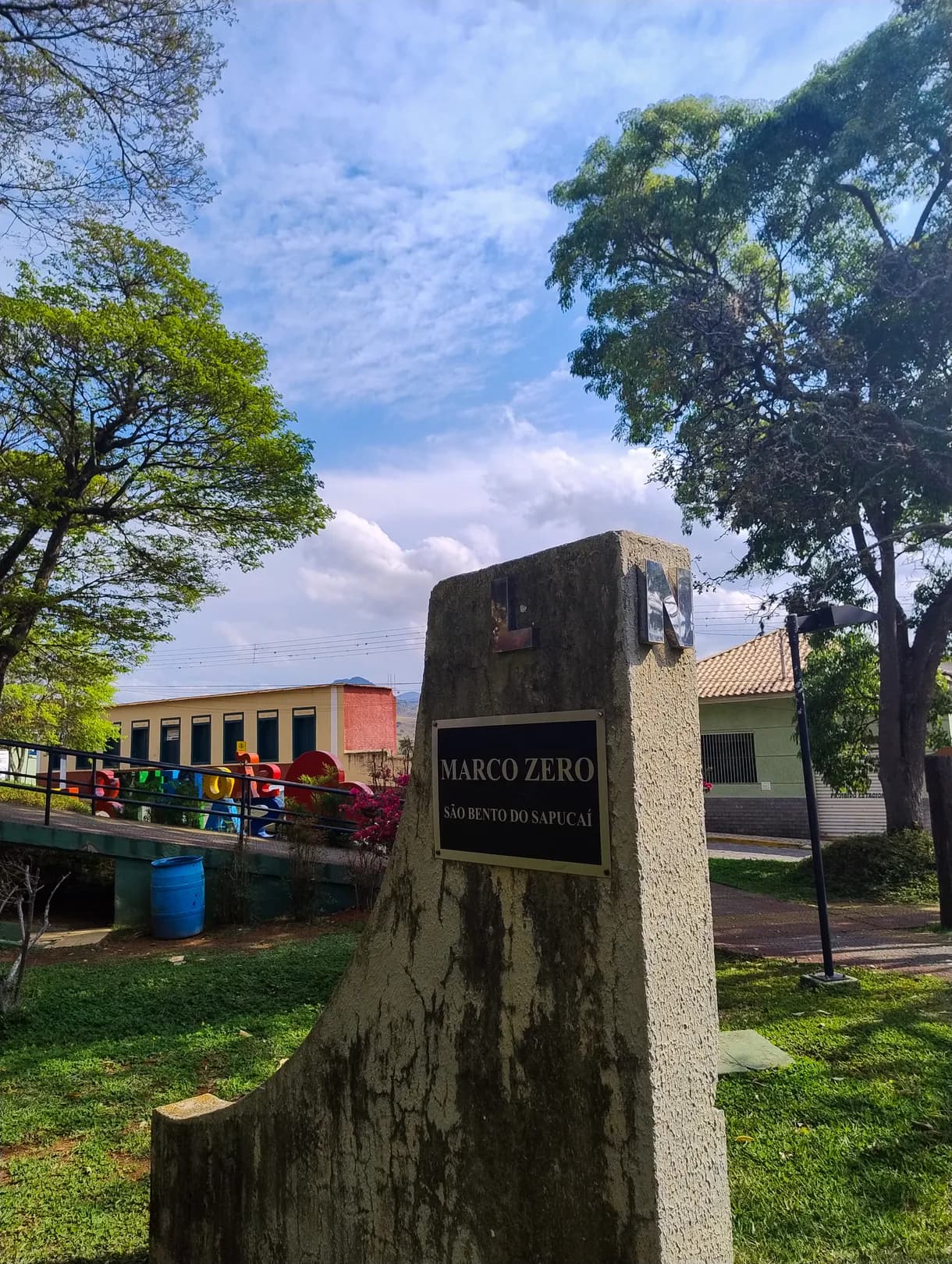 Marco zero de São Bento do Sapucaí na Praça General Marcondes Salgado: bloco com placa no gramado e as letras coloridas da cidade atrás